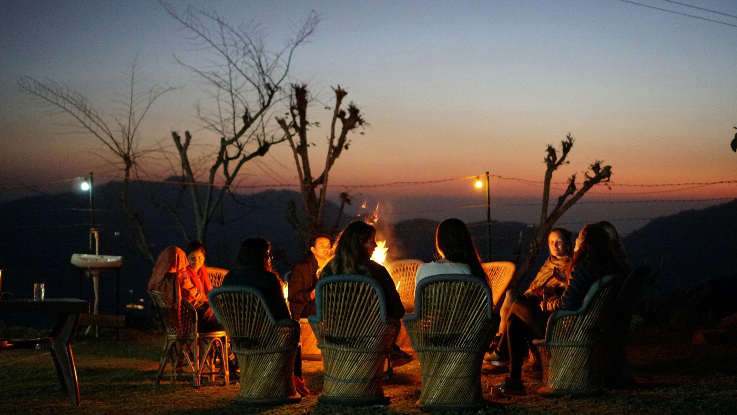 A small fire burning in low evening light beside the shadowed outlines of seated figures — the visual of communal storytelling as an inherited human practice.