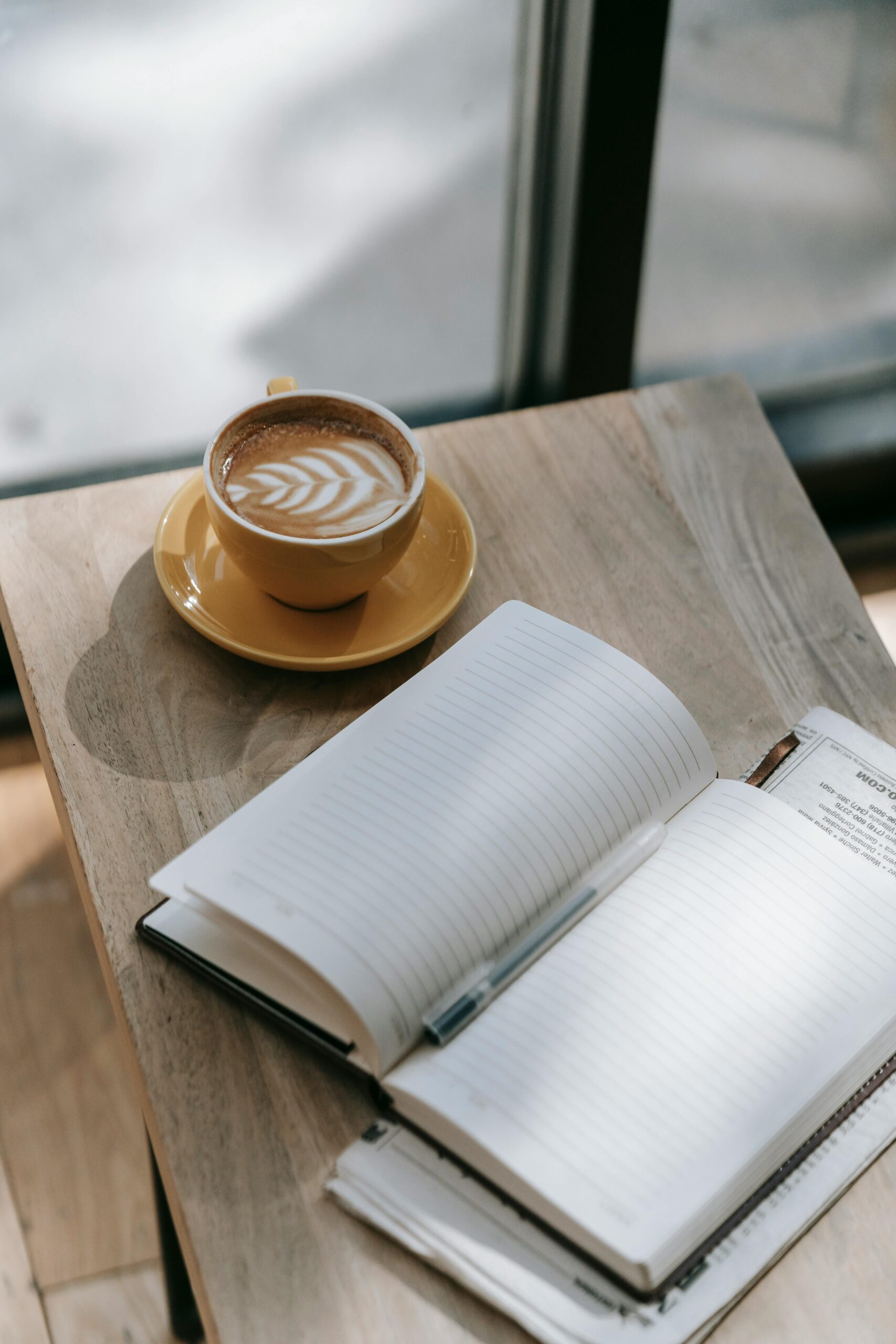 A daybook open beside a steaming cup, sunlight crossing a wooden surface — the visual of a wellness routine designed around the body's own rhythms.